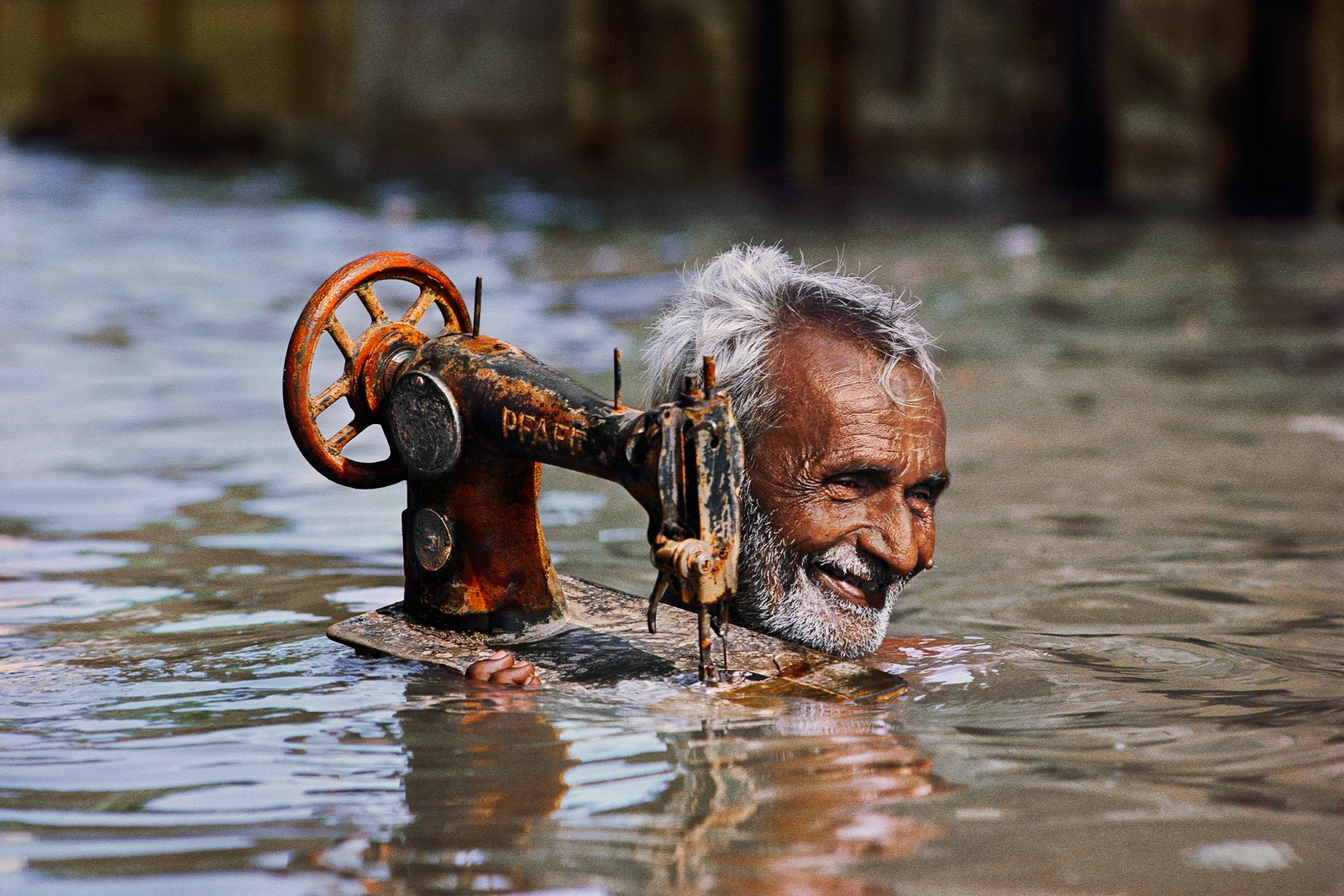 Tailor carries his sewing machine through monsoon floodwaters. Fonte: www.mucsarnok.hu